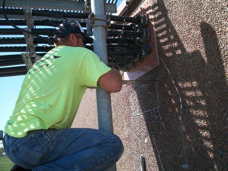 Worker in yellow shirt kneels, working on a pole attached to a building, likely telecommunications equipment.