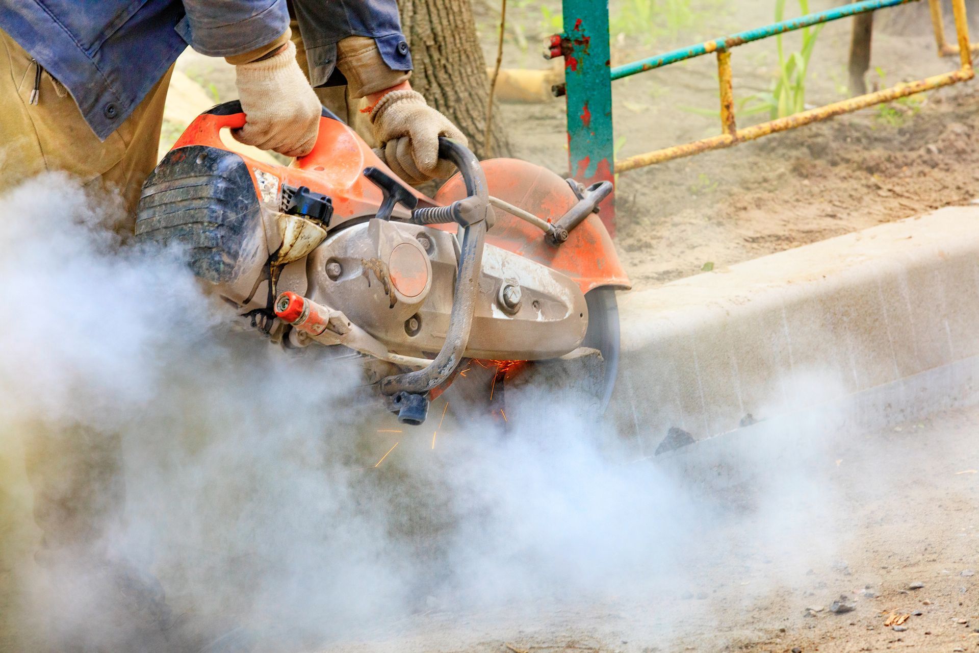 Person wearing gloves uses an orange saw to cut a sidewalk curb, producing dust.