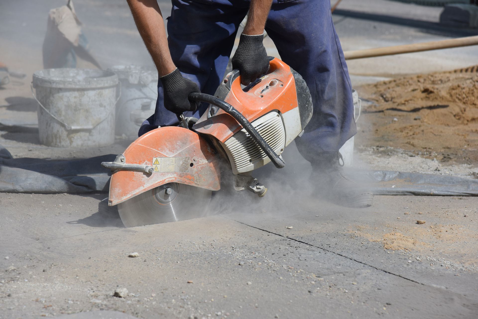 Person in gloves using a concrete saw to cut pavement, dust rising.