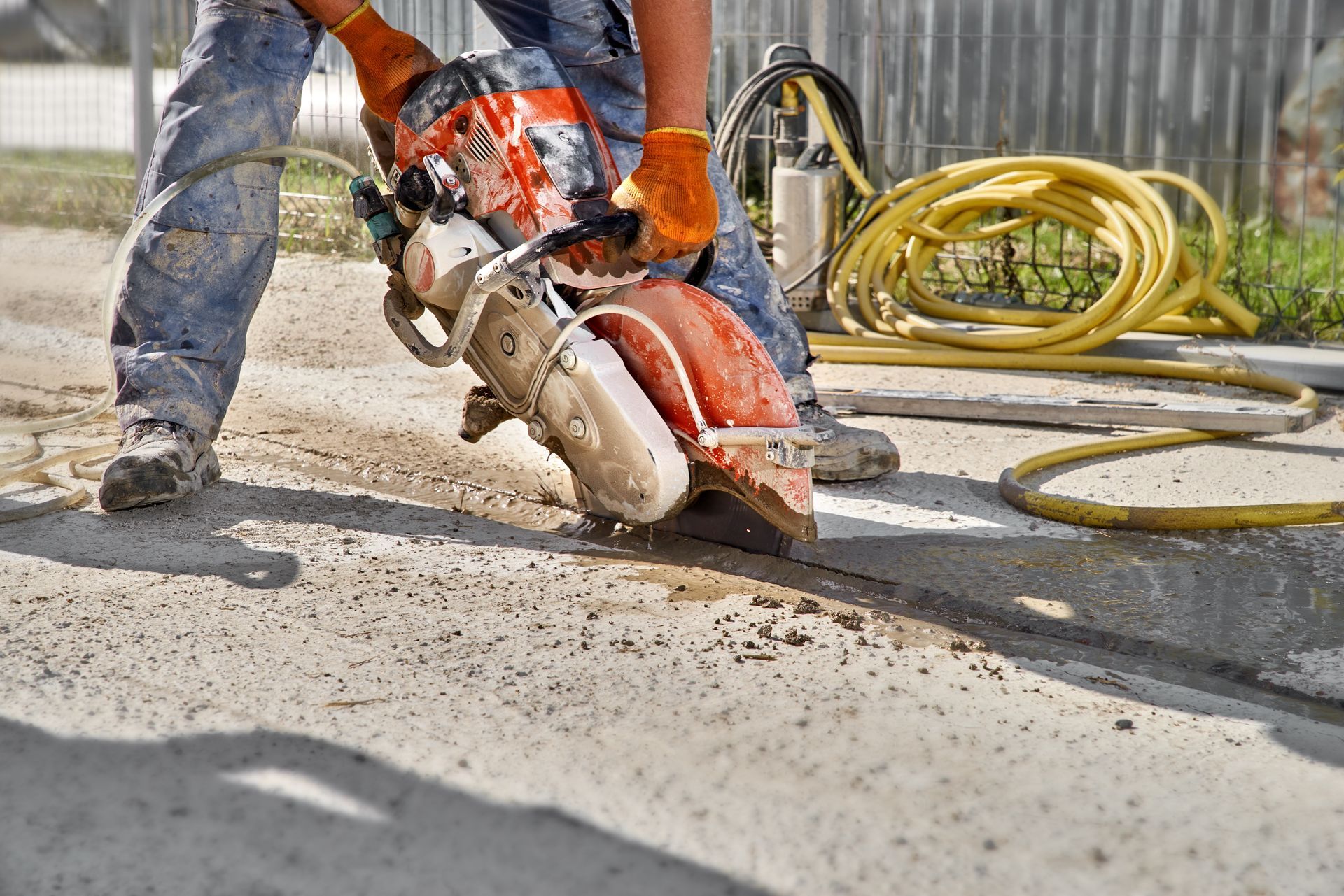 Worker cutting concrete with an orange saw, wearing gloves and jeans.