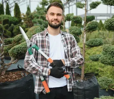Man holding large pruning shears, standing in a greenhouse with potted plants, smiling.