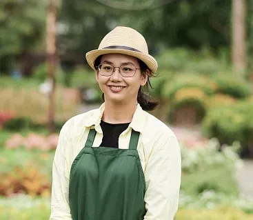Woman wearing glasses, hat, and apron smiles outdoors in a garden setting.
