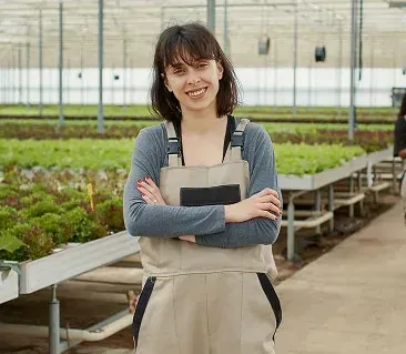 Woman with arms crossed, smiling in a greenhouse with rows of plants.