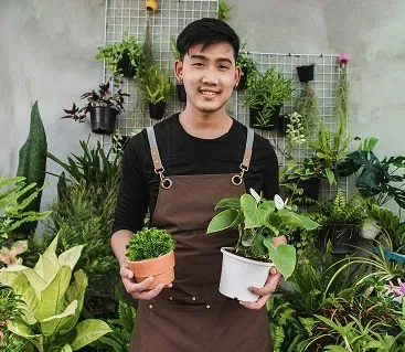 Man in apron smiles, holding potted plants in a lush plant-filled space.