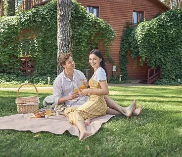 Couple having a picnic on a blanket in front of a wooden cabin covered in greenery.