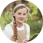 Woman with blonde braid wearing apron holds tablet outdoors.