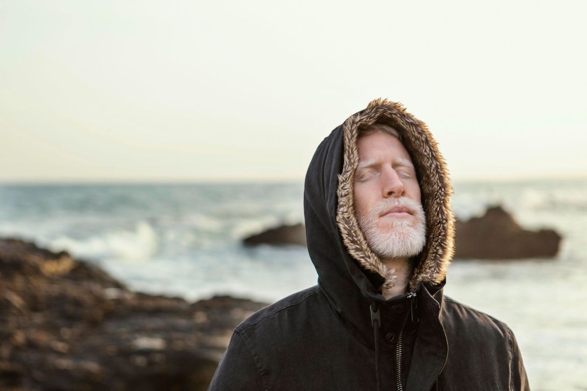 A man with a beard is wearing a hooded jacket on the beach.