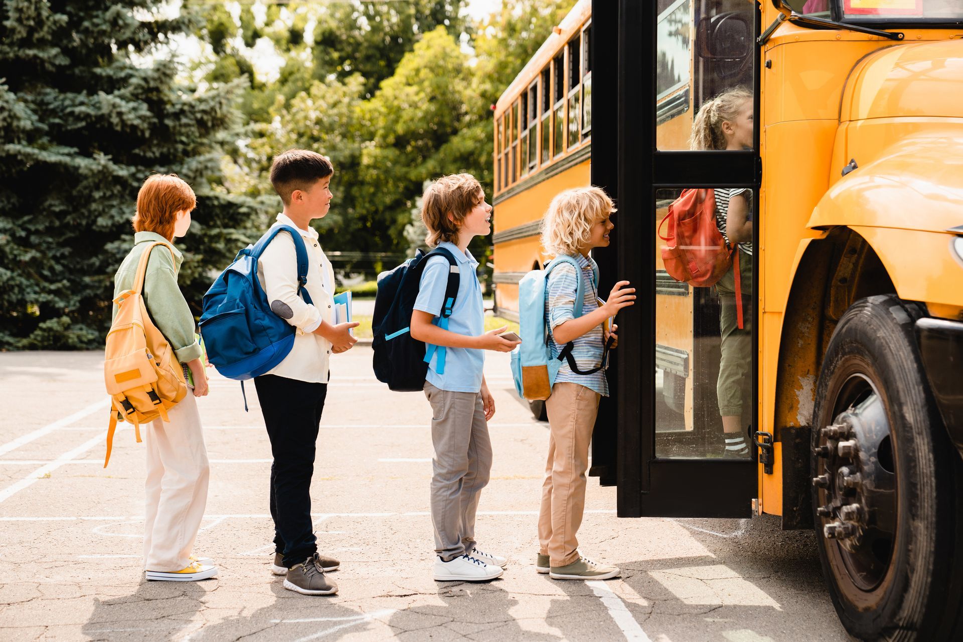 Kids boarding a yellow bus charter for a school trip lined up with backpacks in a sunny parking lot.