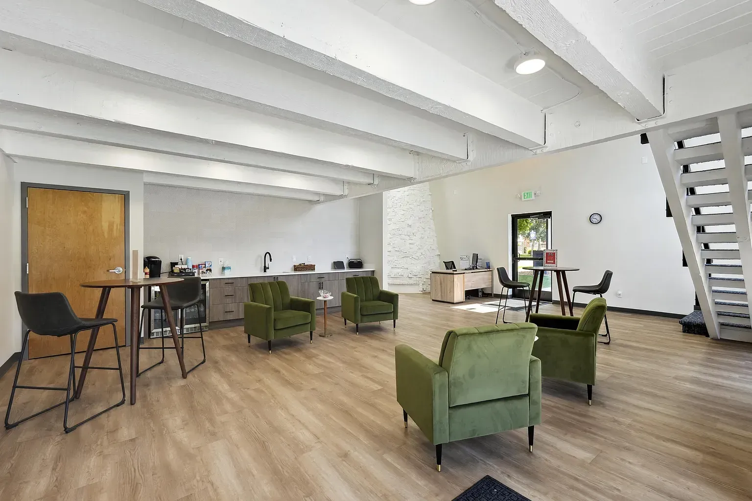 Modern office lounge with white walls, wooden floors, green velvet armchairs, a high-top table, and a staircase.