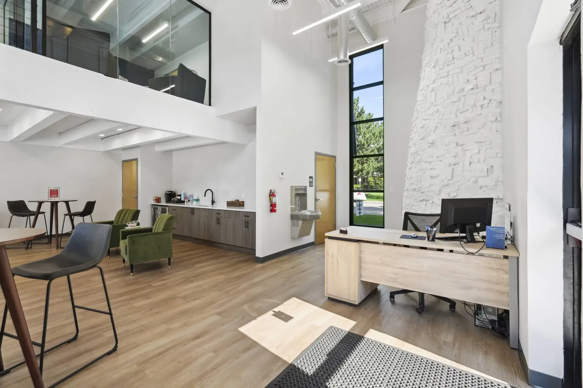Modern office lobby with a reception desk, lounge seating, a kitchenette, and a high ceiling with tall windows.