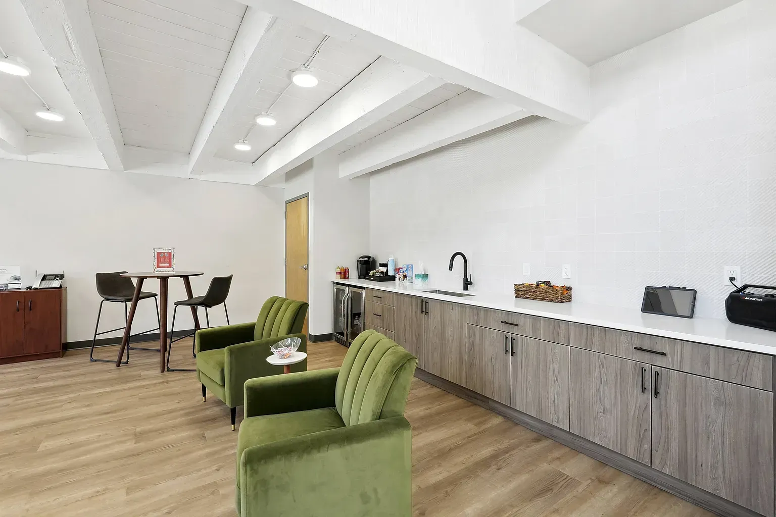A modern breakroom with two green velvet chairs, a wood-toned counter with a sink, and a small bistro table in the corner.