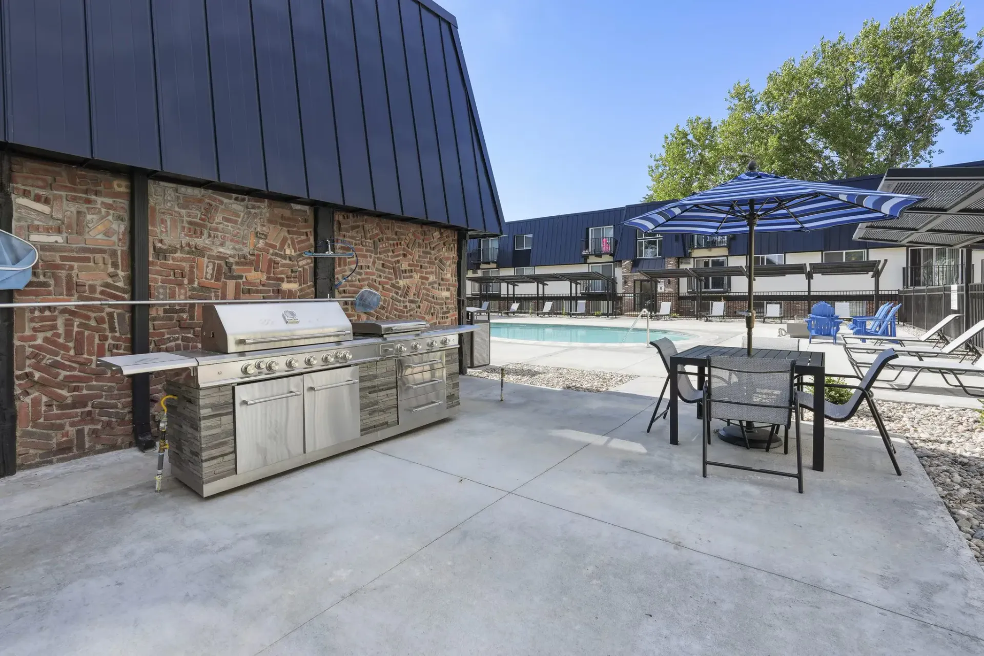 Outdoor poolside patio with a stainless steel grill, a table with chairs under a blue striped umbrella, and brick siding.