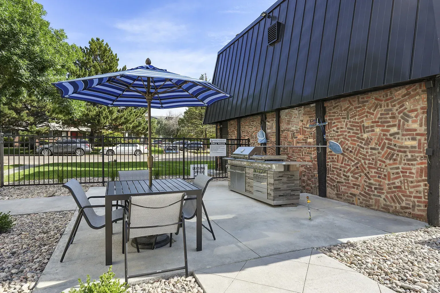 An outdoor patio with a table, blue striped umbrella, and chairs next to a stone building with a metal-paneled roof.