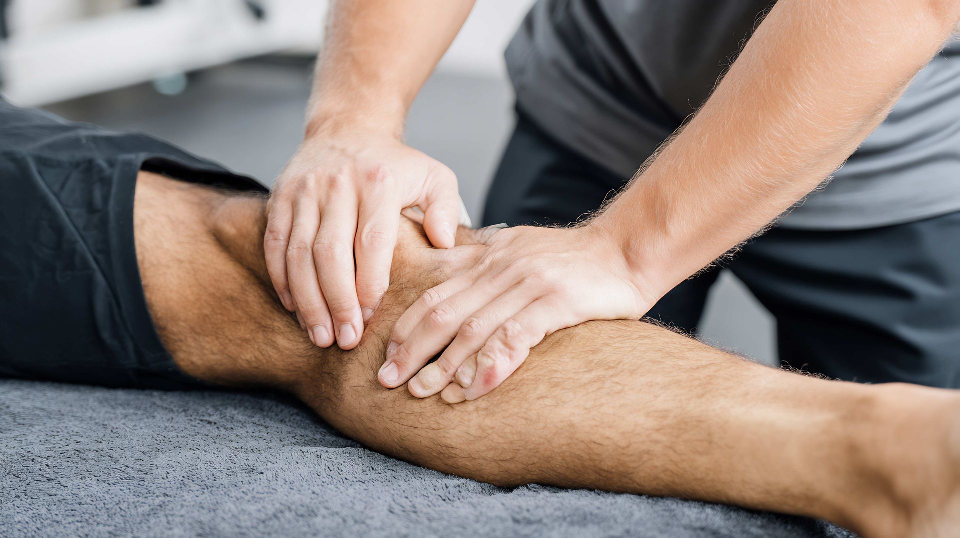Hands massaging a person’s knee on a gray surface, suggesting physical therapy or injury treatment