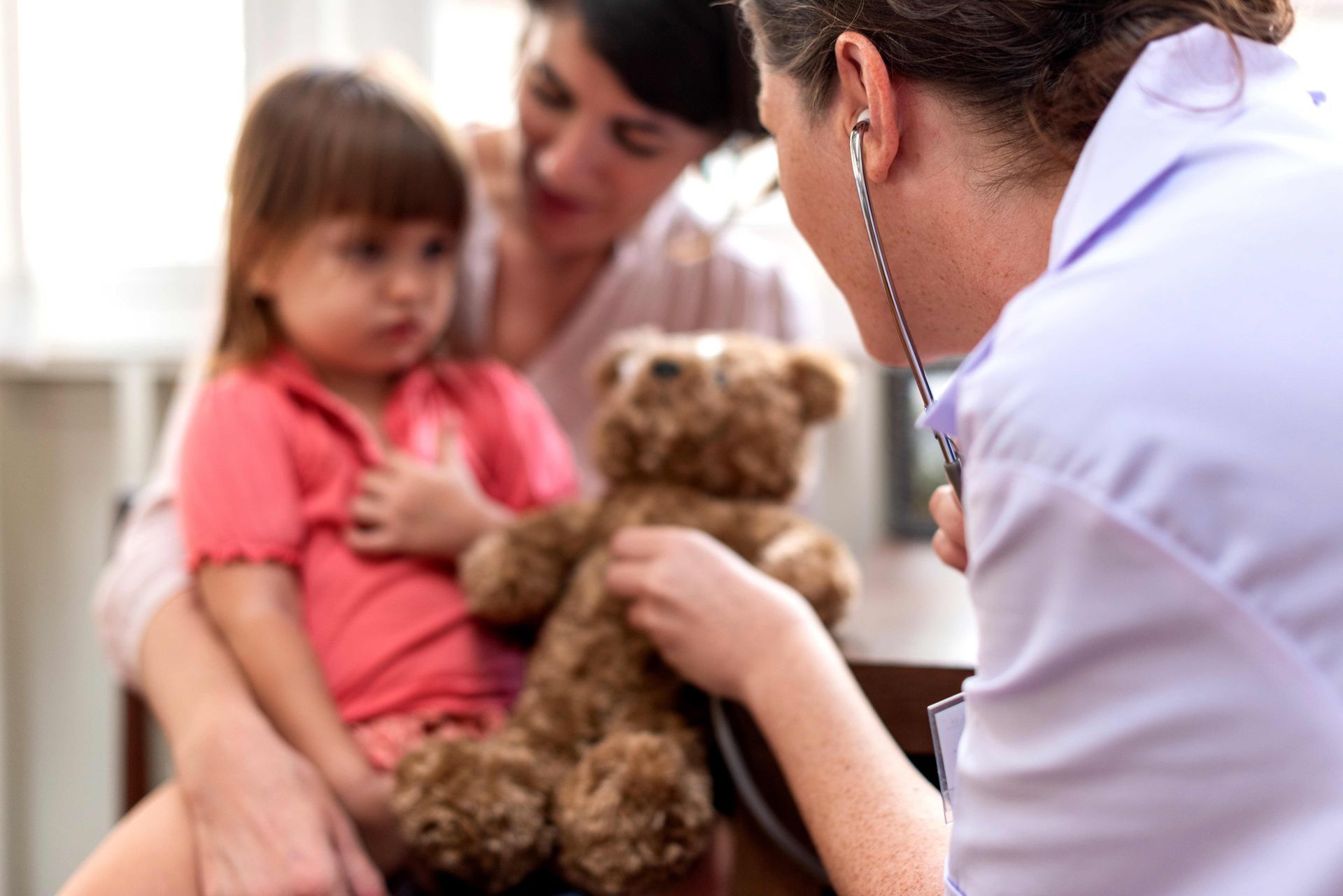 Doctor examining a child holding a teddy bear, with a parent nearby