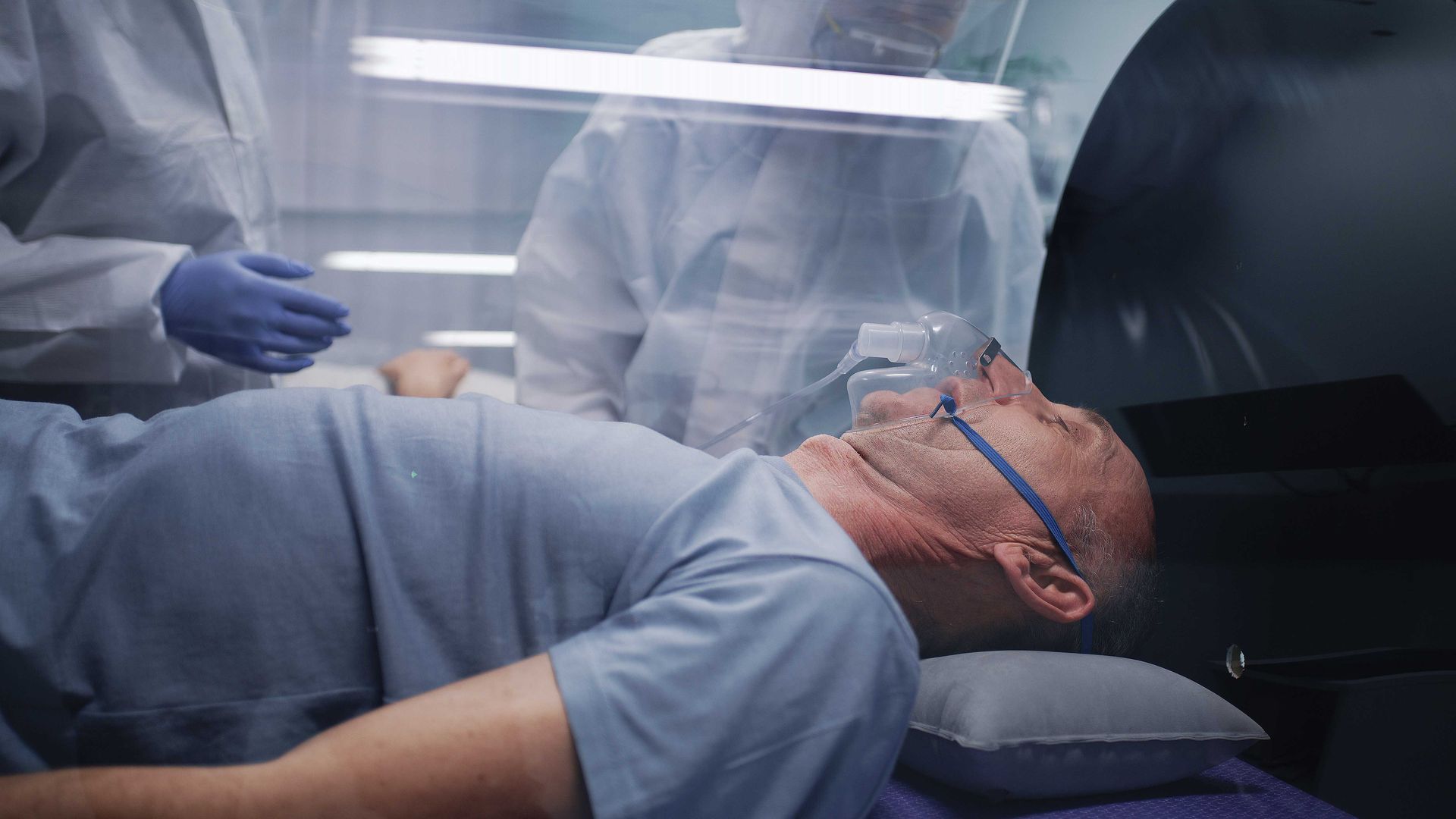 Patient lying on a hospital bed under a clear isolation tent, wearing an oxygen mask.