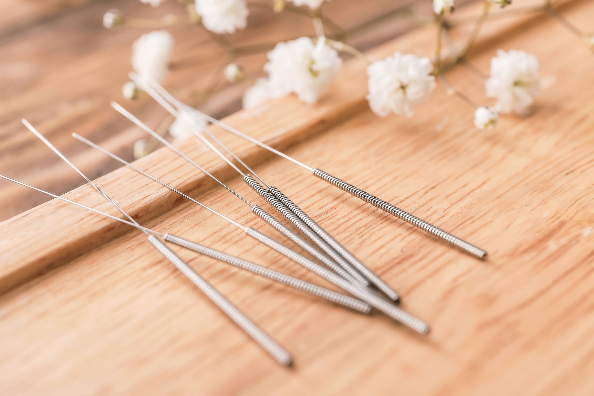 Metal acupuncture needles on a wooden surface with small white flowers in the background