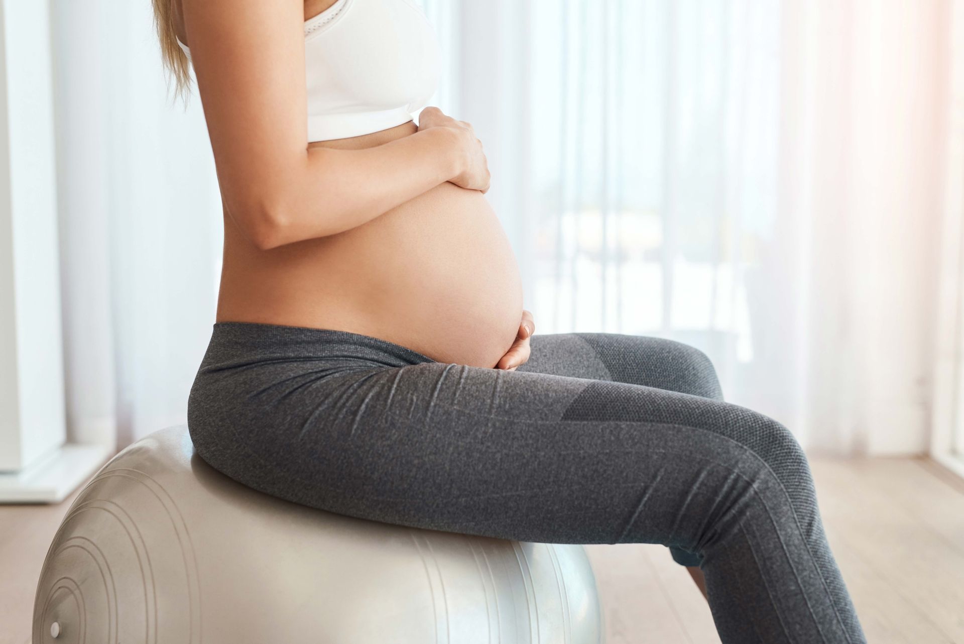 Pregnant person sitting on a white ottoman in a bright room, side profile with belly visible Pregnant person sitting on a white ottoman in a bright room, side profile with belly visible