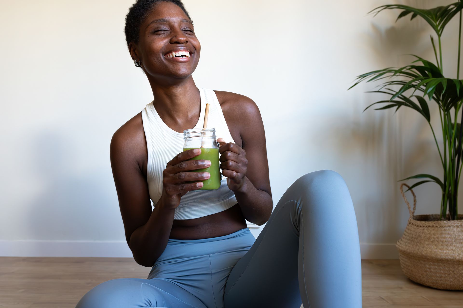Smiling person in workout clothes holding a green smoothie on a yoga mat beside a potted plant
