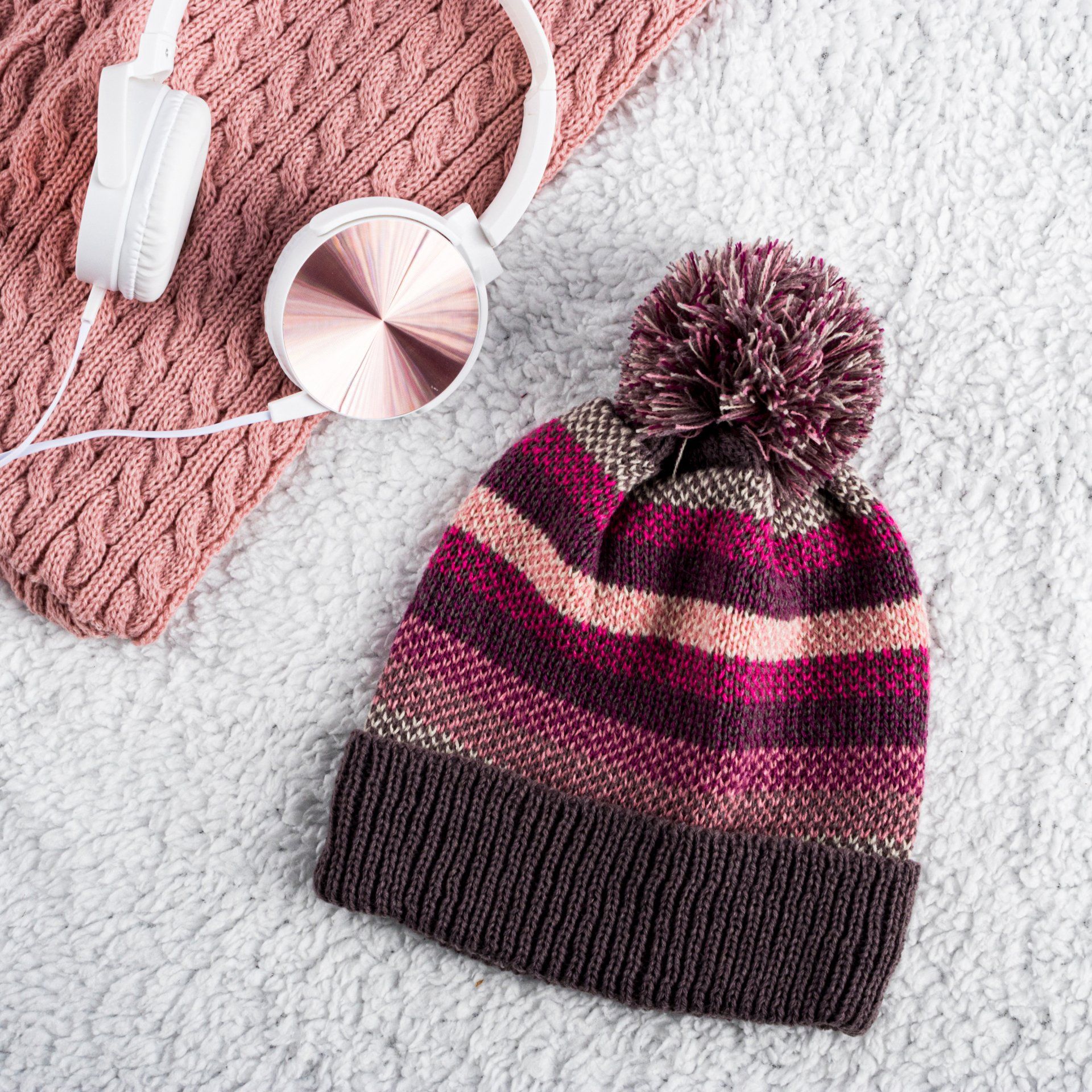 A knitted hat with a pom pom is sitting on a blanket next to headphones and a scarf.