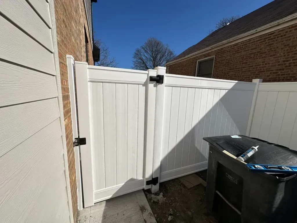 White vinyl gate in a narrow side yard beside brick house and trash bins under bright sun