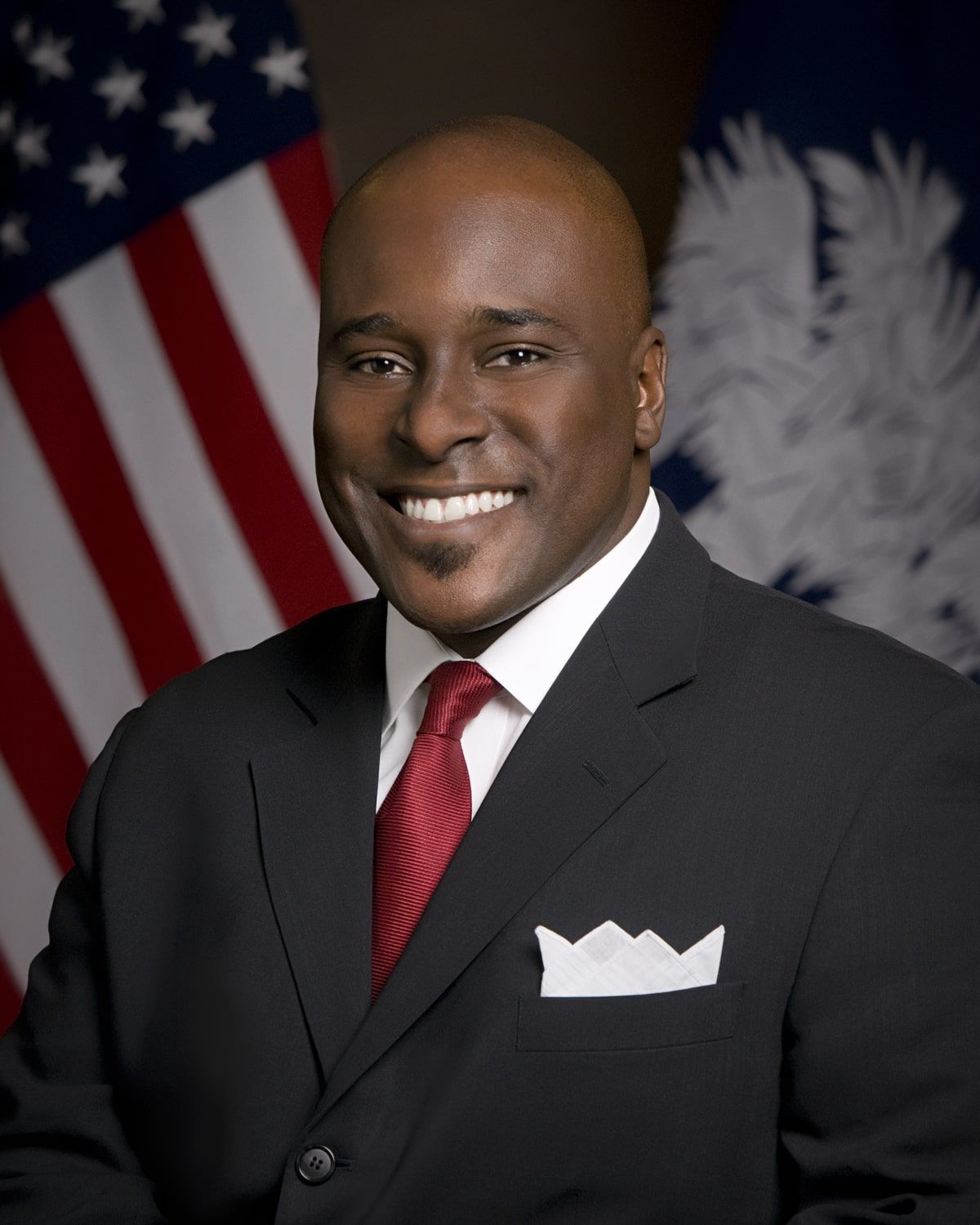 Man in suit smiling, with flags of the United States and Louisiana in the background.