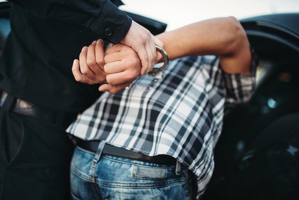 A person in a plaid shirt is being handcuffed by a uniformed officer near a car.