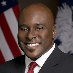 Man in suit with red tie smiling, with US flag and Louisiana seal in background.
