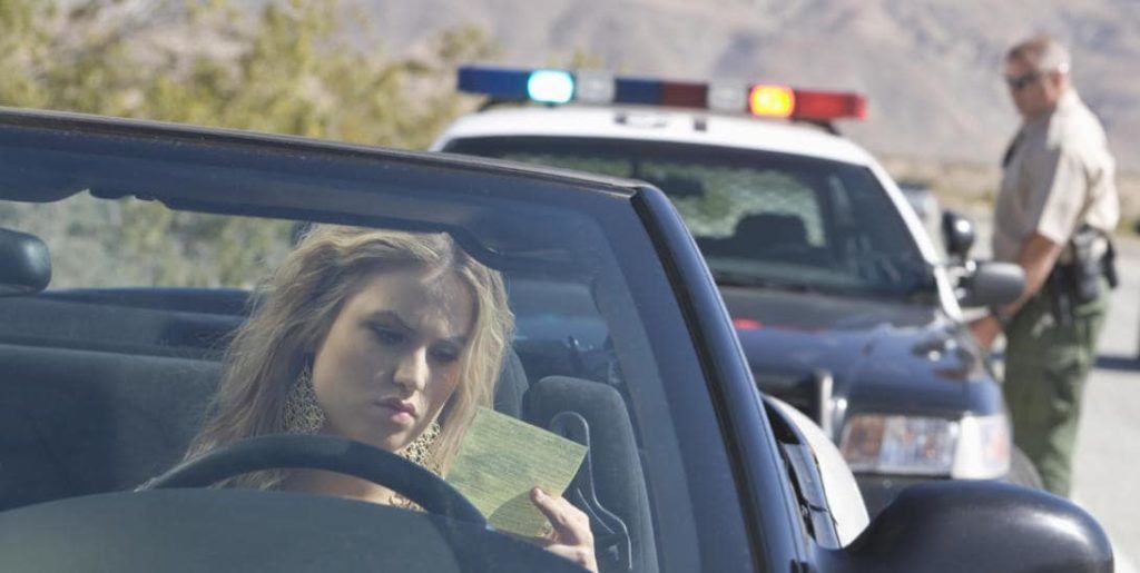 A woman in a convertible looks at a ticket as a police officer stands nearby his car in a desert setting.