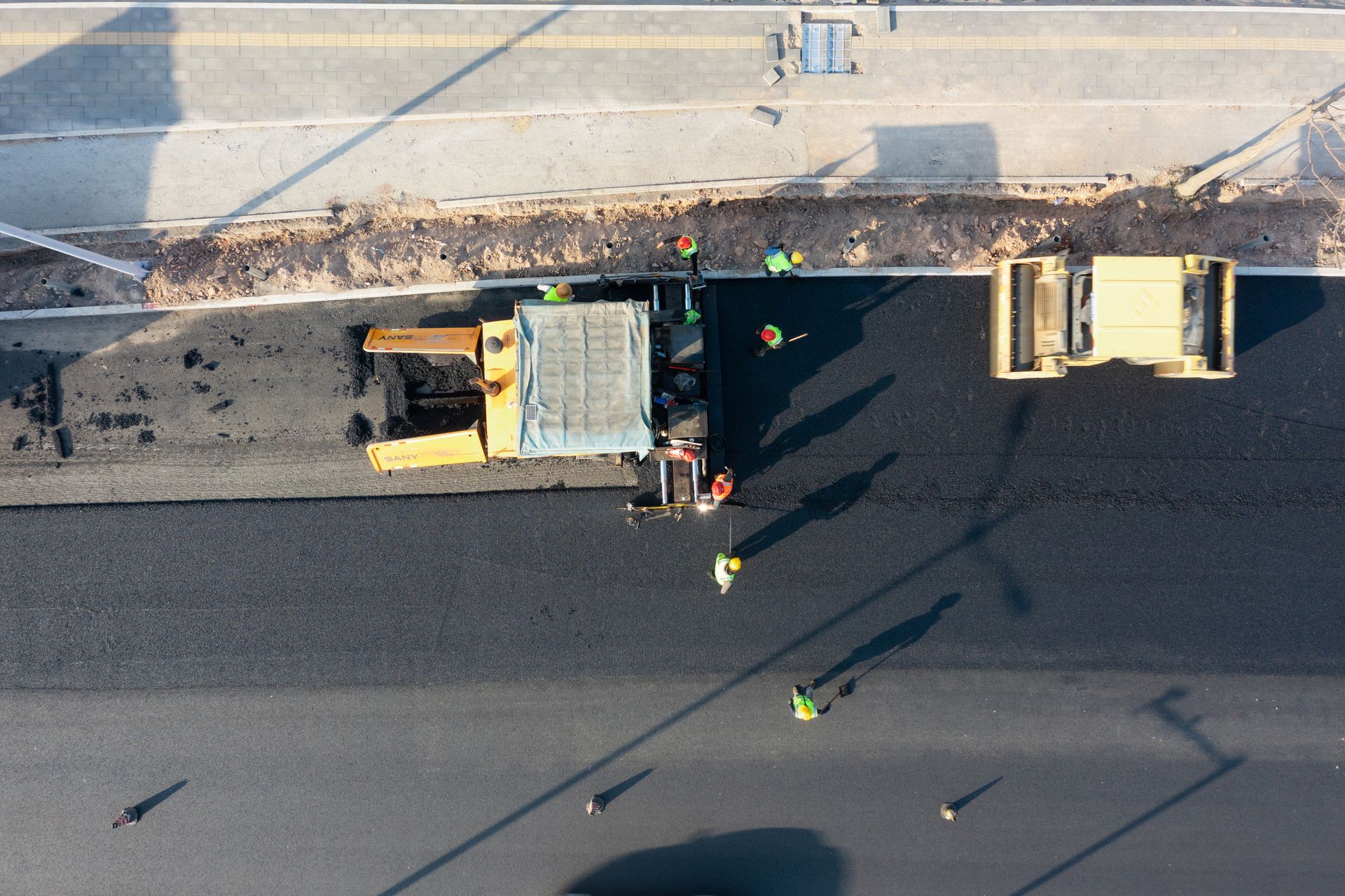 Road construction: Asphalt paver laying fresh pavement, workers in safety vests; next to a sidewalk.