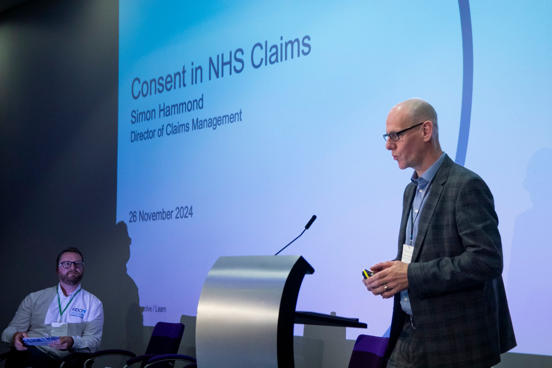 A man is standing at a podium giving a presentation on consent in nhs claims.