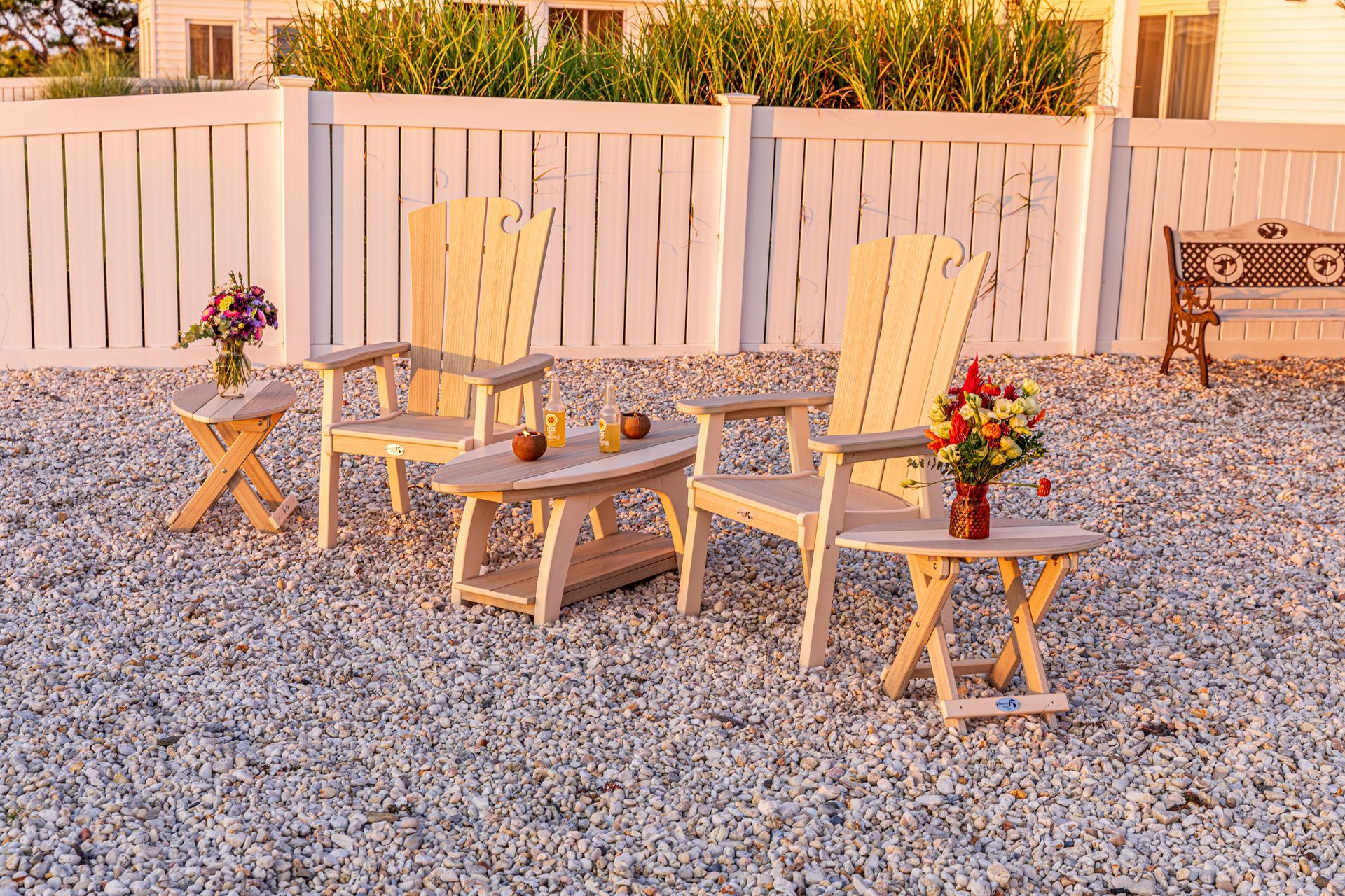 A group of wooden chairs and tables are sitting on gravel in front of a white fence.