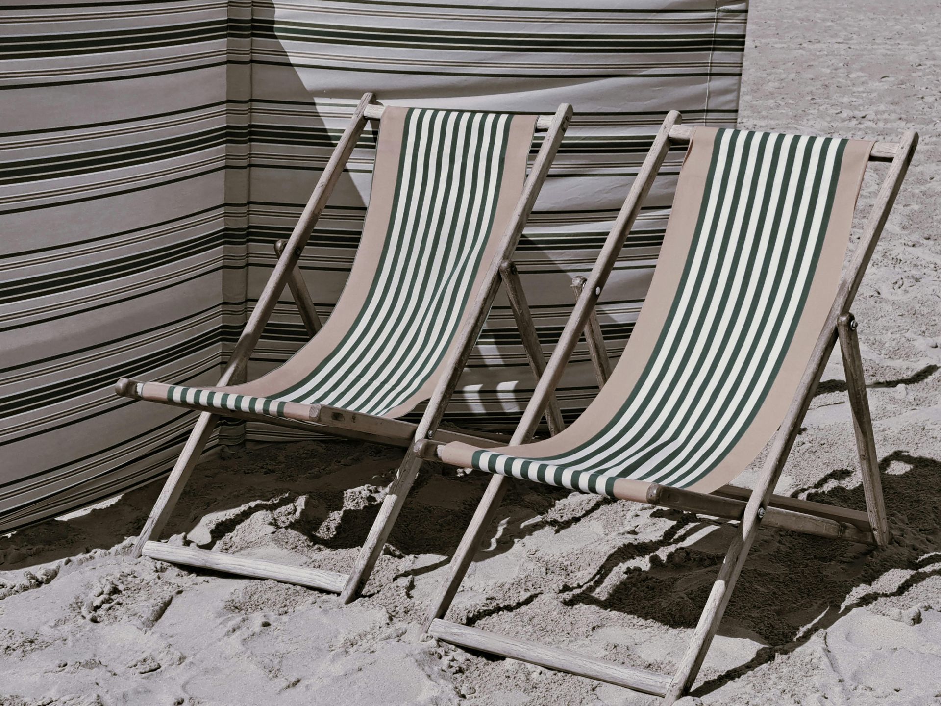 Two striped beach chairs on sand. A striped windbreak is in the background.
