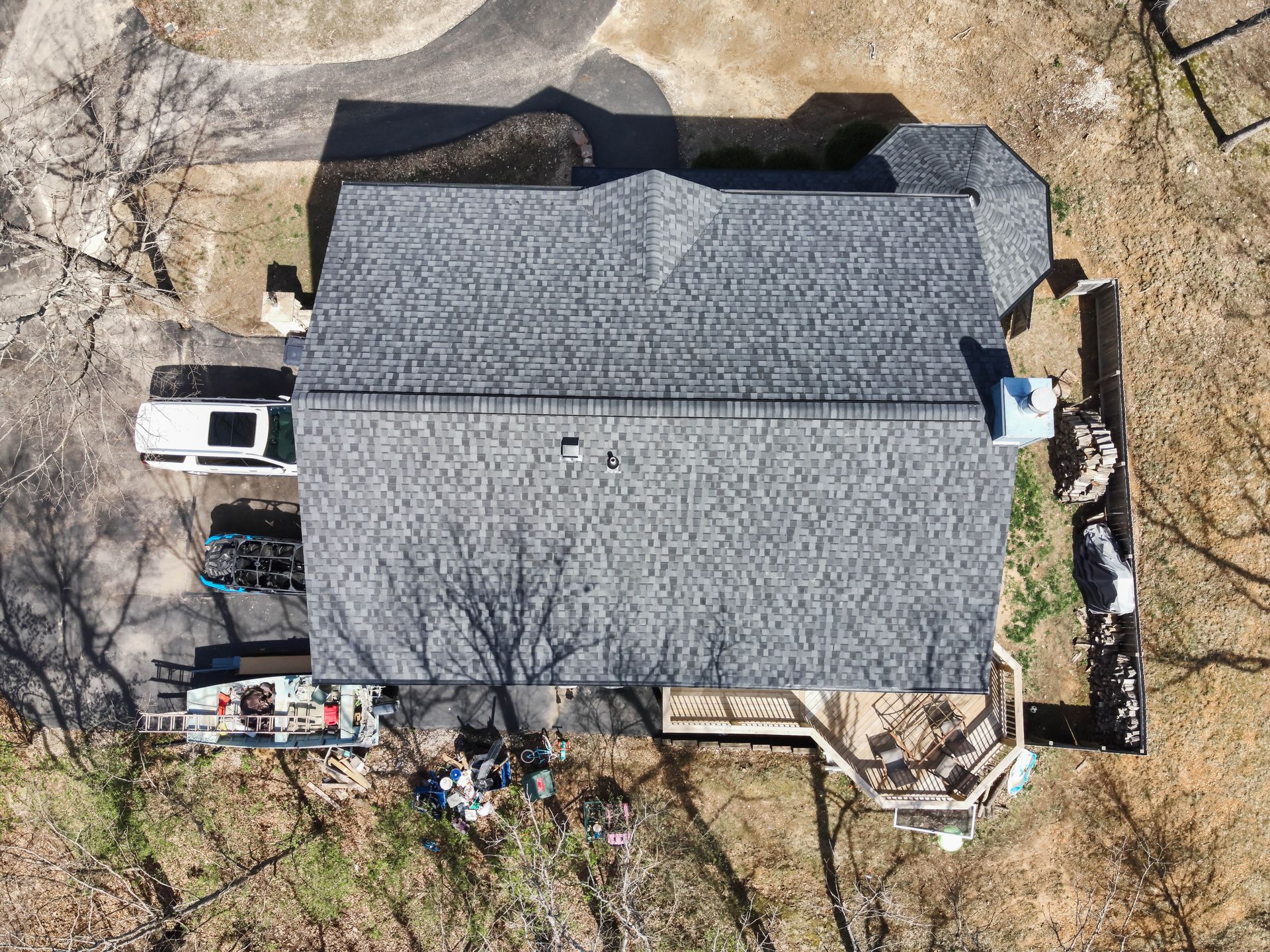 Overhead view of a house with a gray shingled roof, surrounded by trees and parked vehicles in a wooded area.