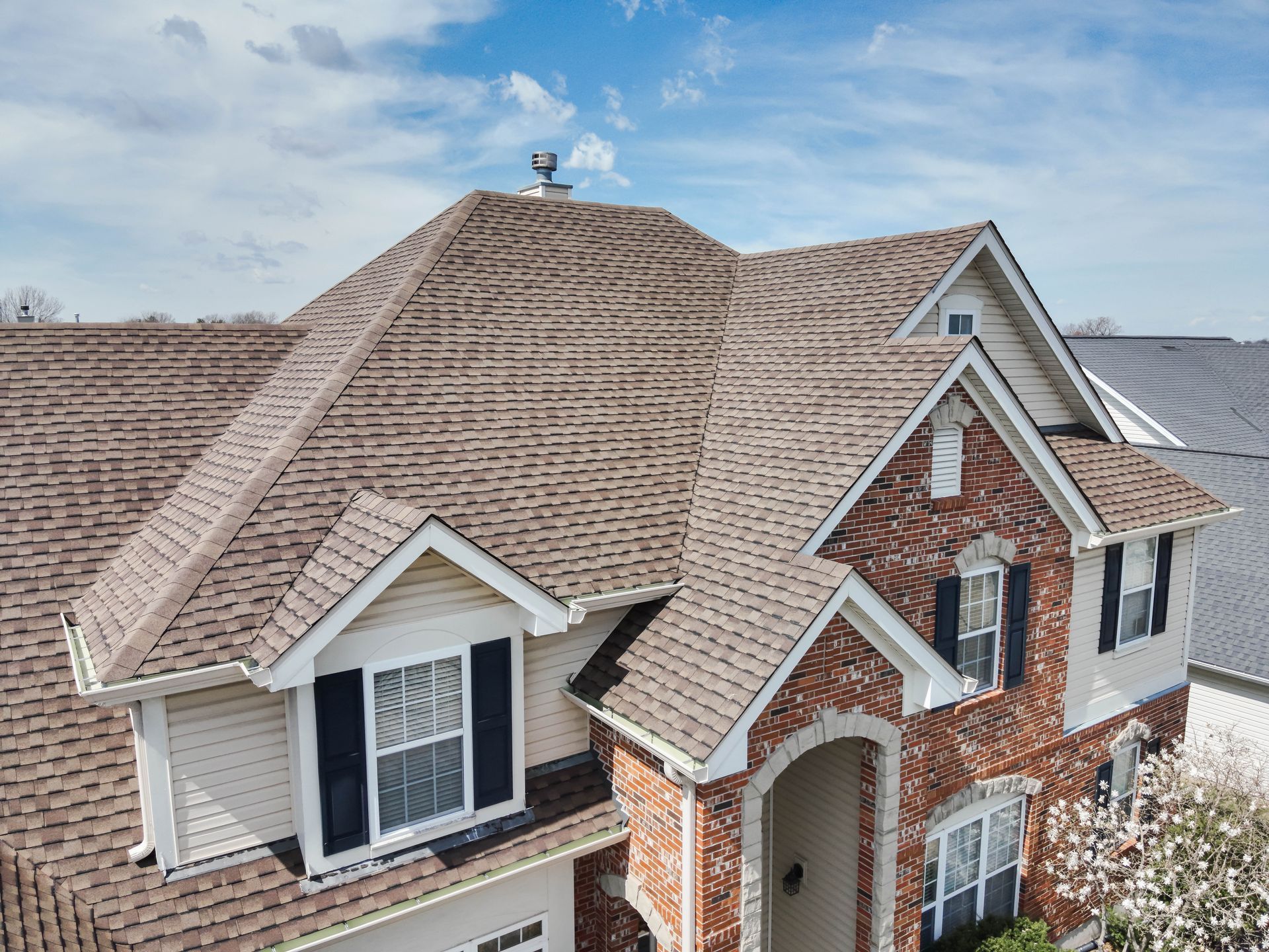 Two-story house with a brown roof, brick facade, and white siding against a blue sky.