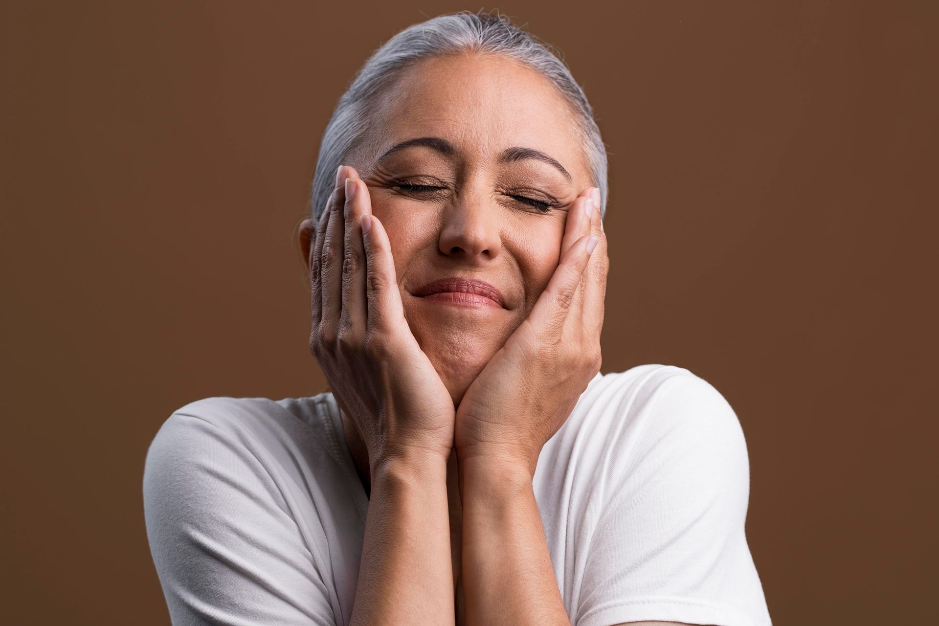 An older woman is smiling with her hands on her face.