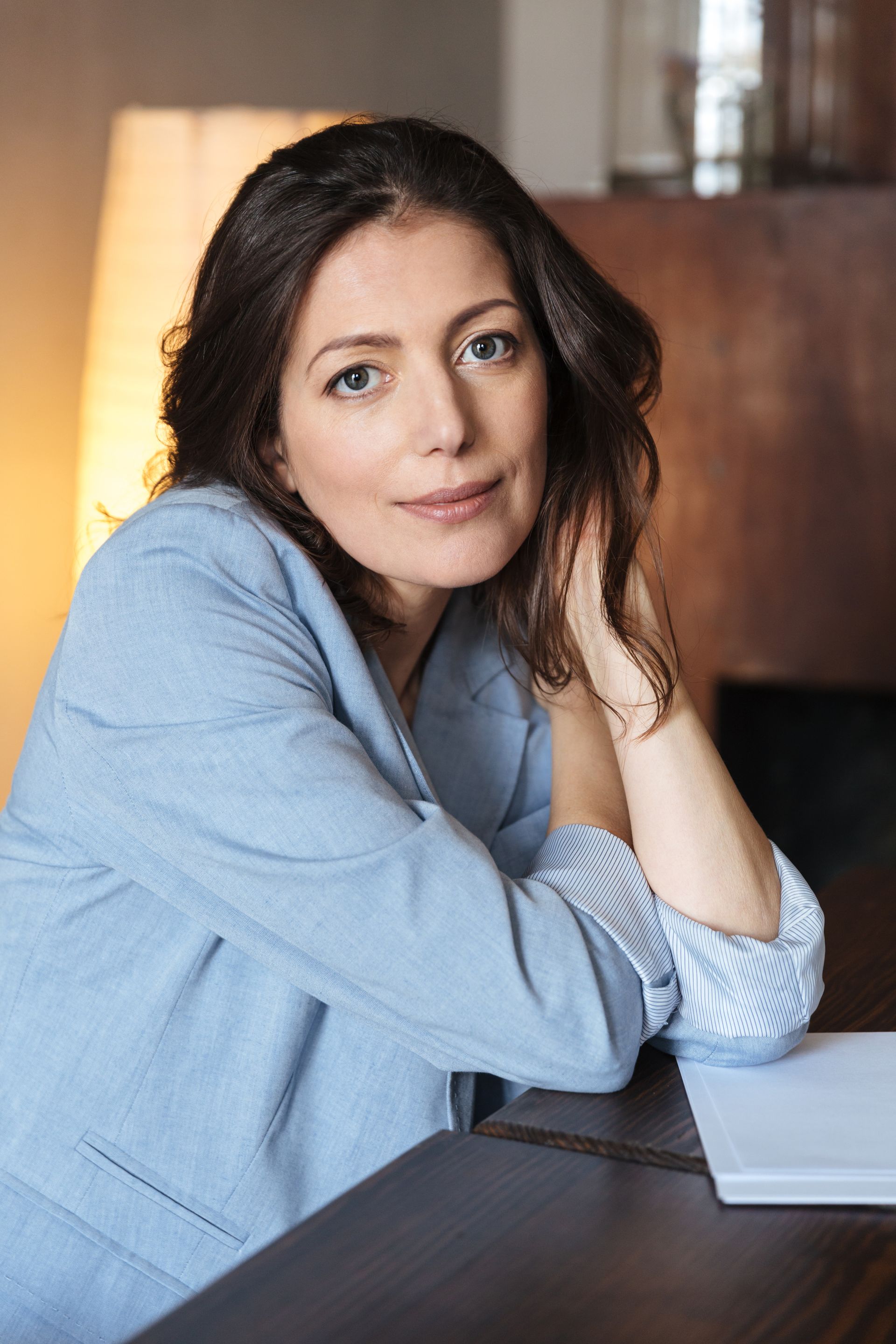 A woman in a blue jacket is sitting at a table with her hands on her chin.