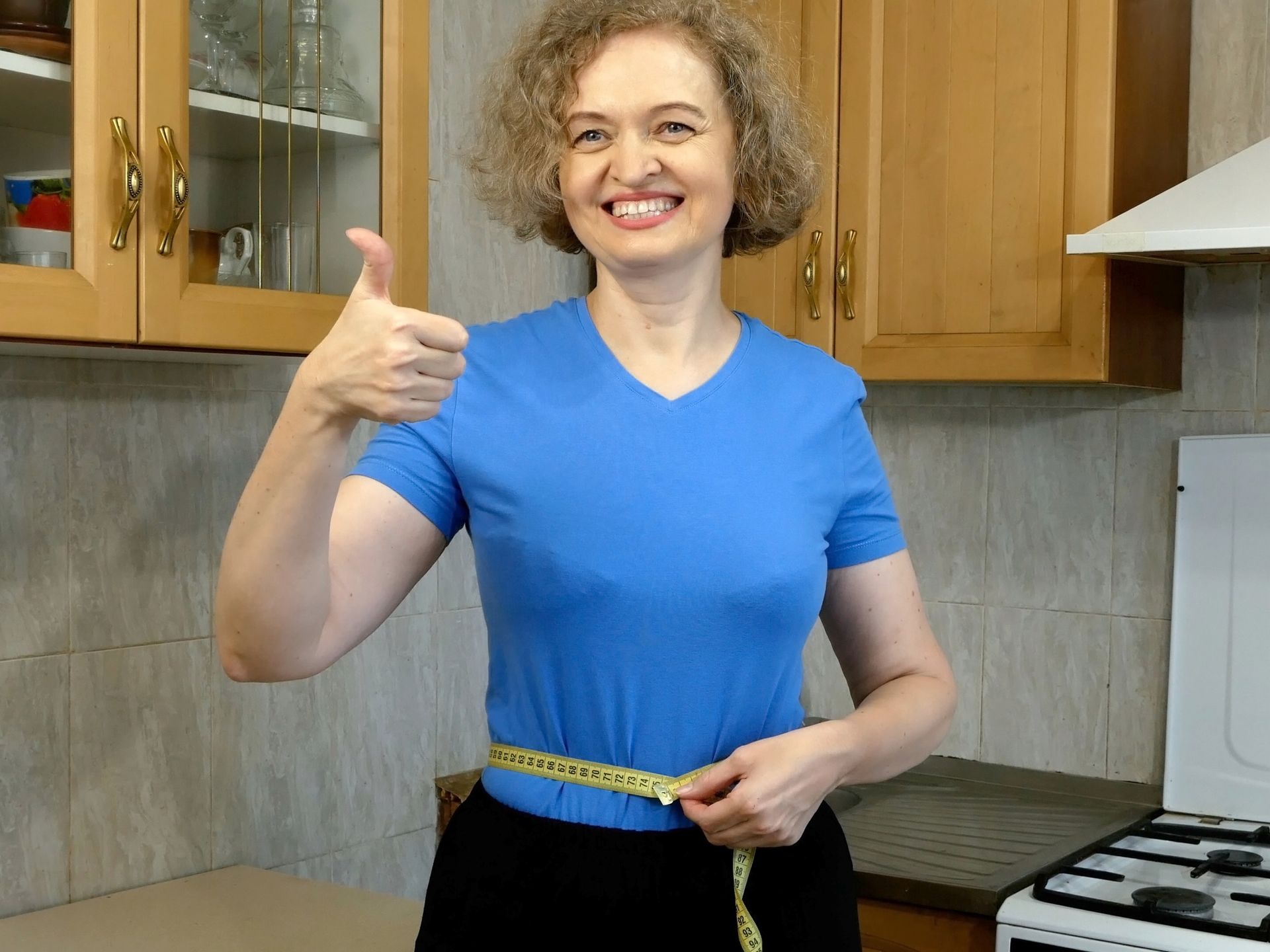 A woman in a blue shirt is measuring her waist and giving a thumbs up