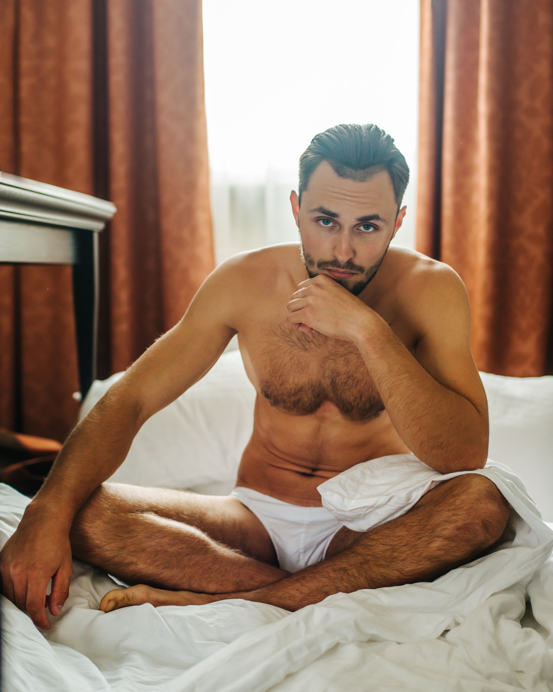 A shirtless man is sitting on a bed with his legs crossed.