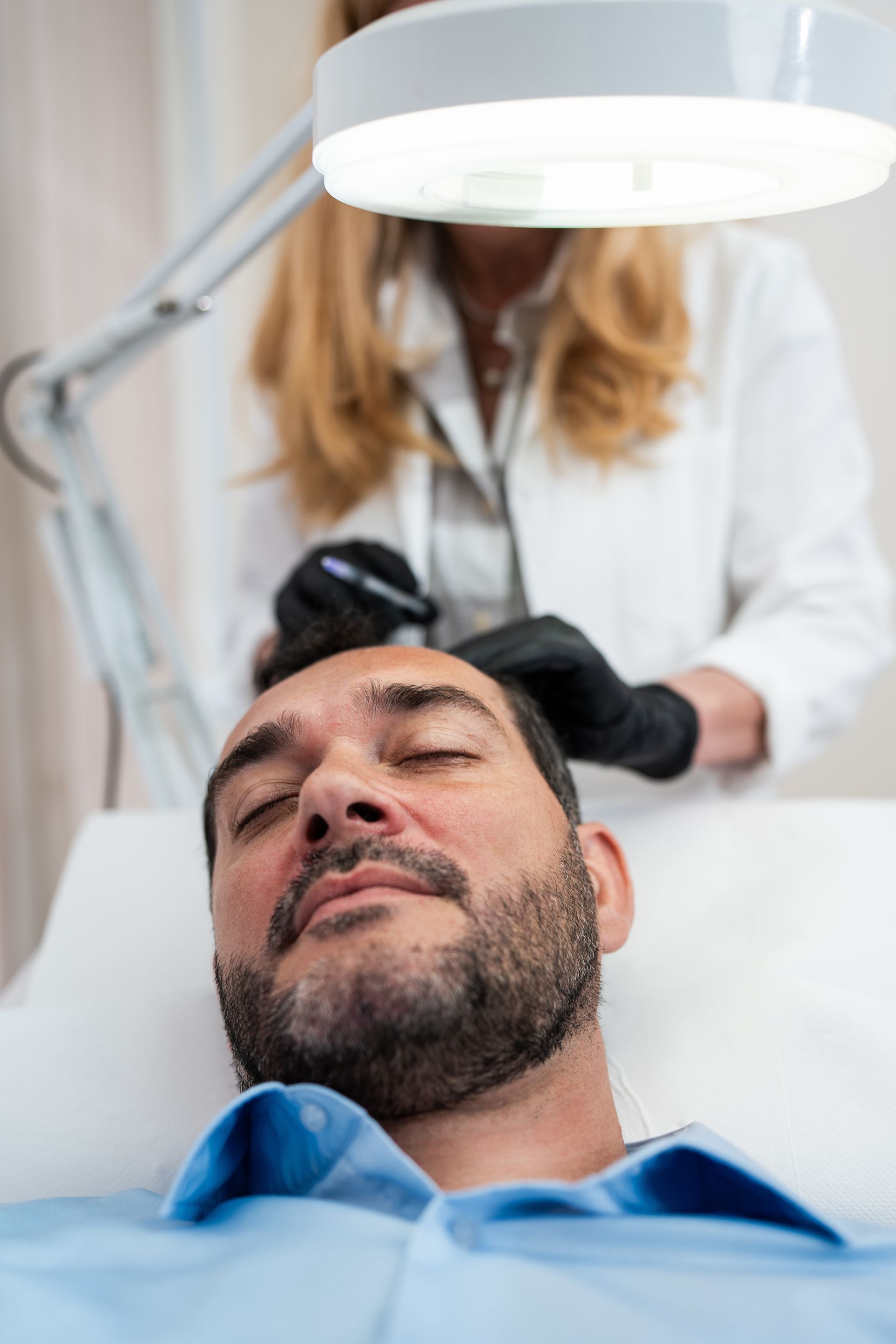 A man with a beard is getting a hair transplant from a female doctor.