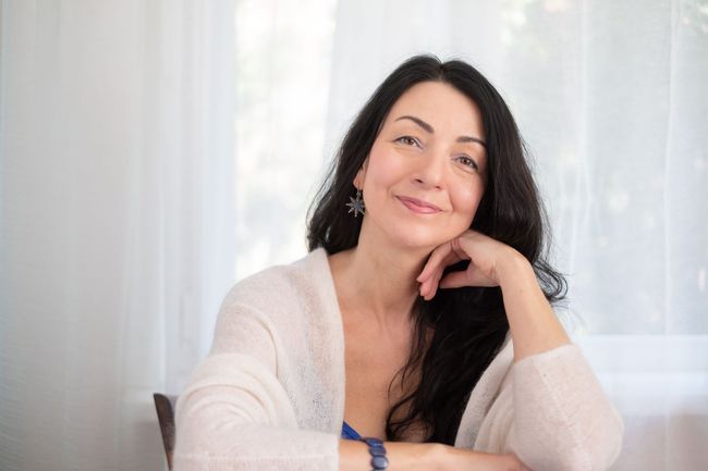 A woman is sitting at a table with her hand on her chin and smiling.