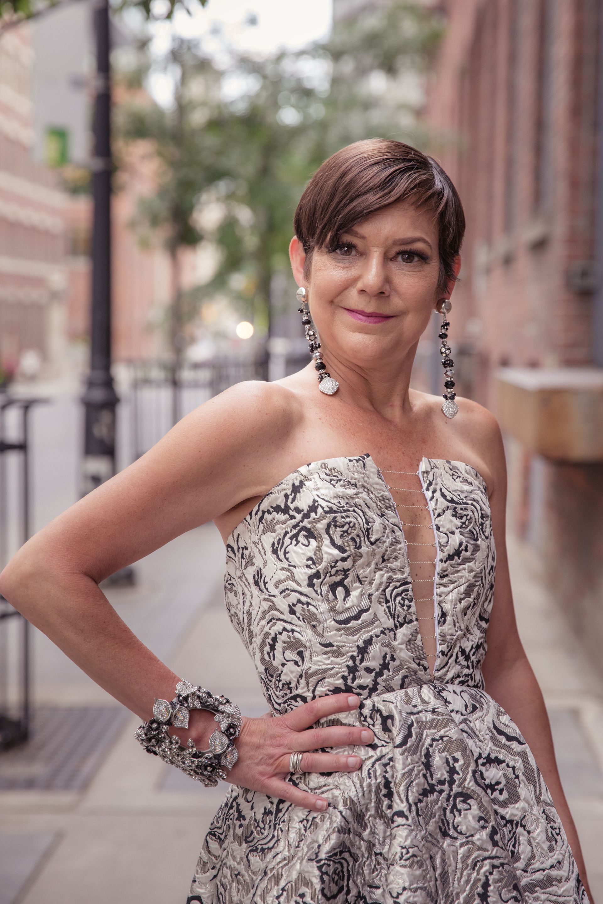 A woman in a strapless dress is standing on a sidewalk with her hand on her hip.