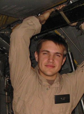 A young man in a military uniform is standing in a dark room with his arms over his head.