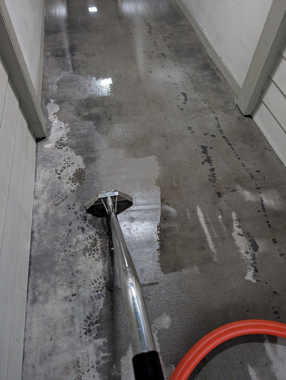 A person cleaning a concrete floor with a wet vacuum. Narrow hallway with white walls.