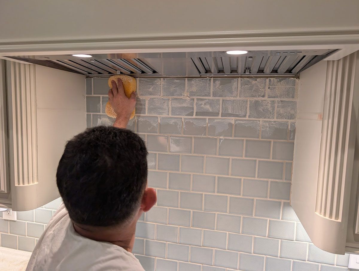 Person sponges blue tile backsplash under a range hood in a kitchen.