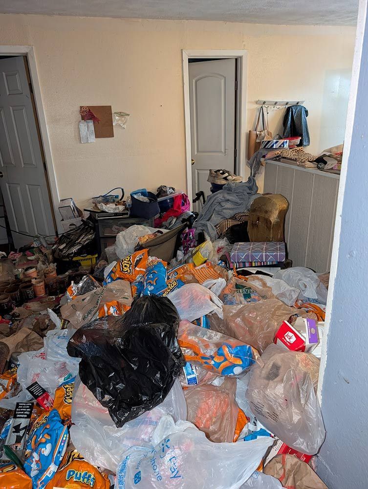Room cluttered with garbage bags and food packaging; a door, closet and clothing rack in the background.