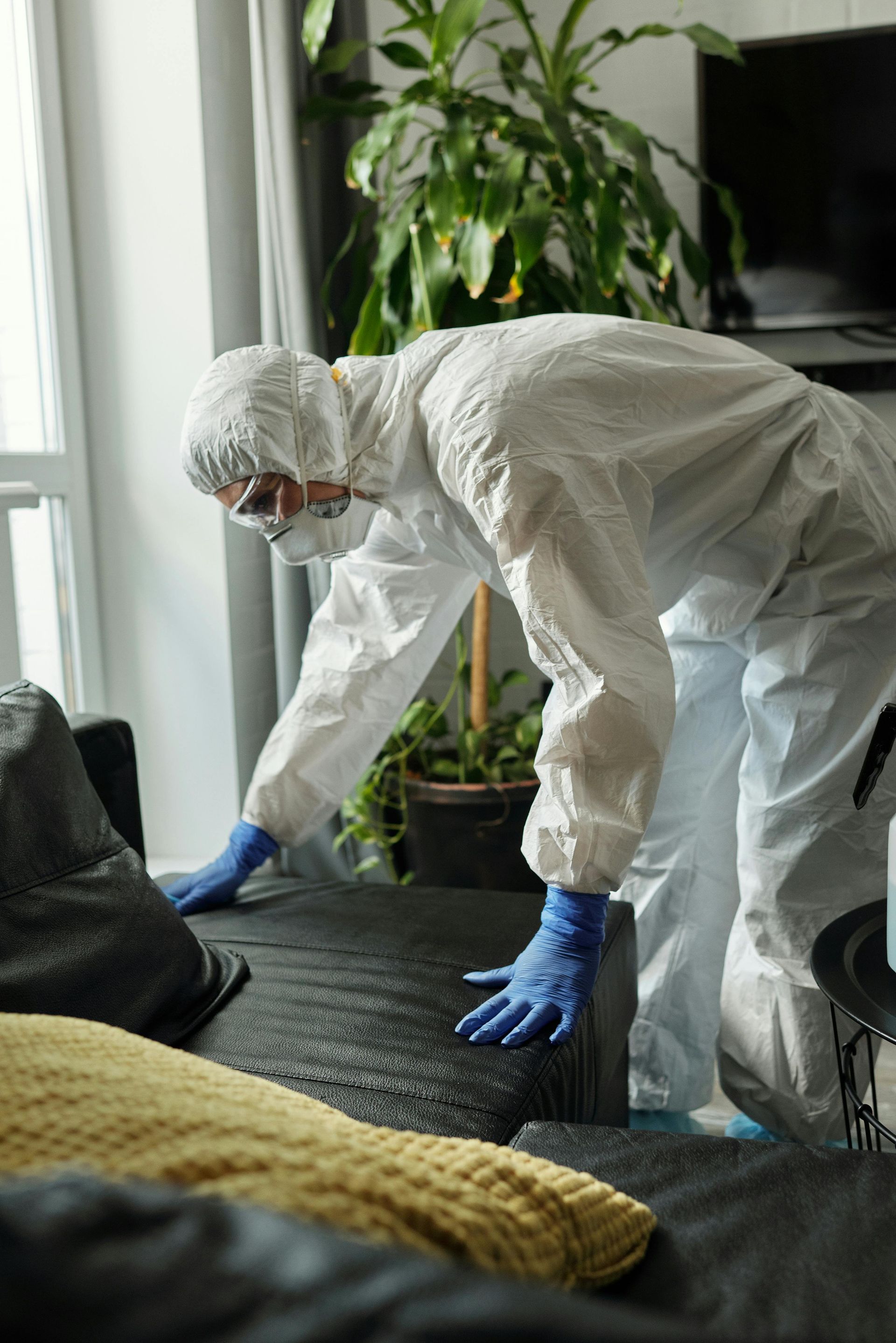A man in a protective suit is cleaning a couch in a living room.