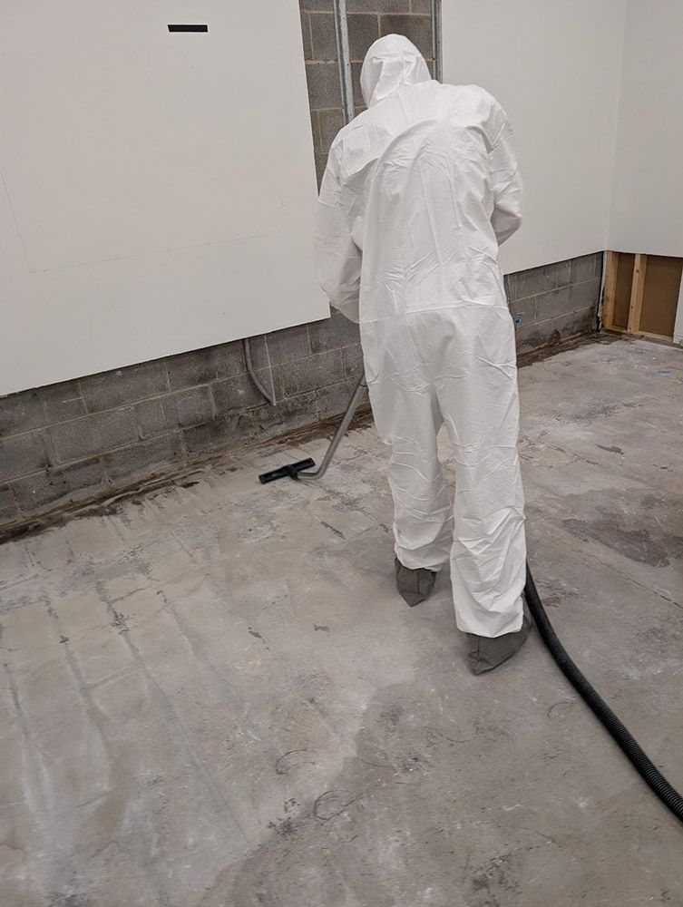 Person in a white protective suit vacuuming a concrete floor in a room with exposed cinder block wall.