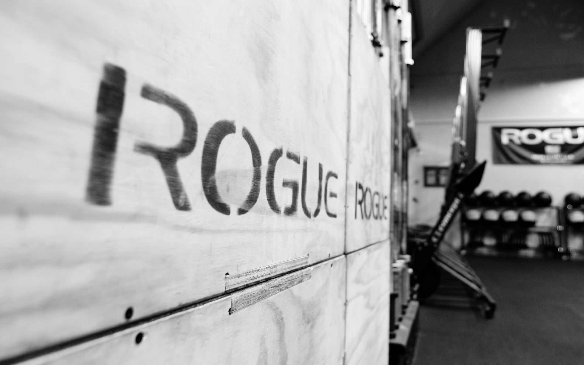 Das Logo „Rogue“ ist auf eine Holzwand in einem Fitnessstudio aufgesprüht; im Hintergrund sind Hanteln und andere Geräte zu sehen.