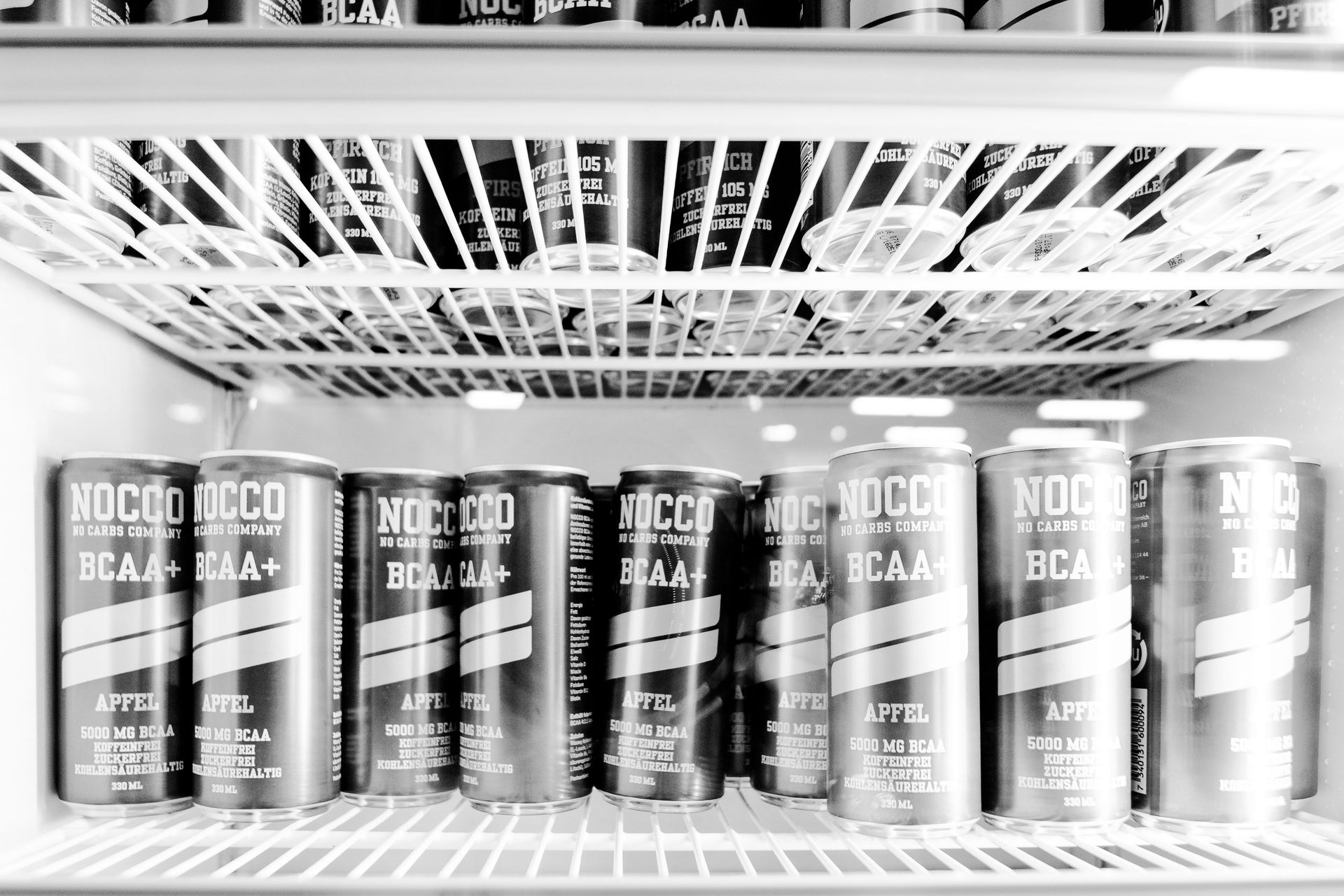 Ein Kühlschrankregal voller Dosen des Energy-Drinks „NOCCO“, aufgenommen in Schwarz-Weiß.